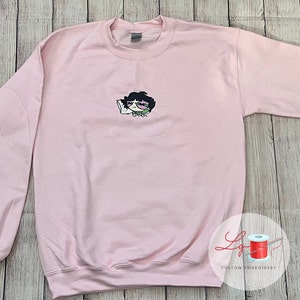 May include: A pink sweatshirt with an embroidered cartoon character wearing glasses and holding a phone. The character has black hair and is wearing a green shirt.