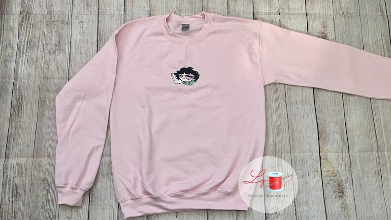 May include: A pink sweatshirt with an embroidered cartoon character wearing glasses and holding a phone. The character has black hair and is wearing a green shirt.
