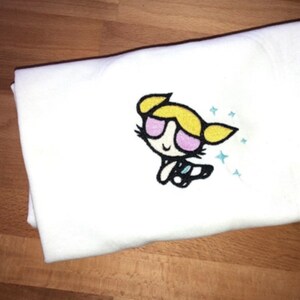 May include: White t-shirt with an embroidered image of a cartoon character with blonde hair, pink sunglasses, and a blue and white outfit. The character is surrounded by blue stars.