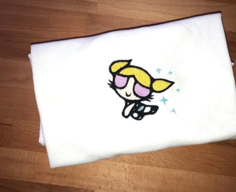 May include: White t-shirt with an embroidered image of a cartoon character with blonde hair, pink sunglasses, and a blue and white outfit. The character is surrounded by blue stars.