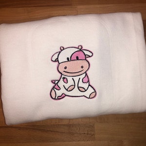 May include: White baby blanket with a pink and white embroidered cow design.