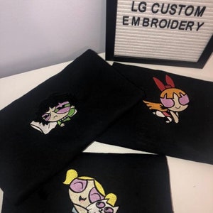 May include: Three black t-shirts with embroidered images of the Powerpuff Girls. The first shirt features Bubbles holding a white cat. The second shirt features Buttercup with a green and purple outfit. The third shirt features Blossom with a red and orange outfit. The shirts are displayed on a white surface with a sign that reads "LG CUSTOM EMBROIDERY".