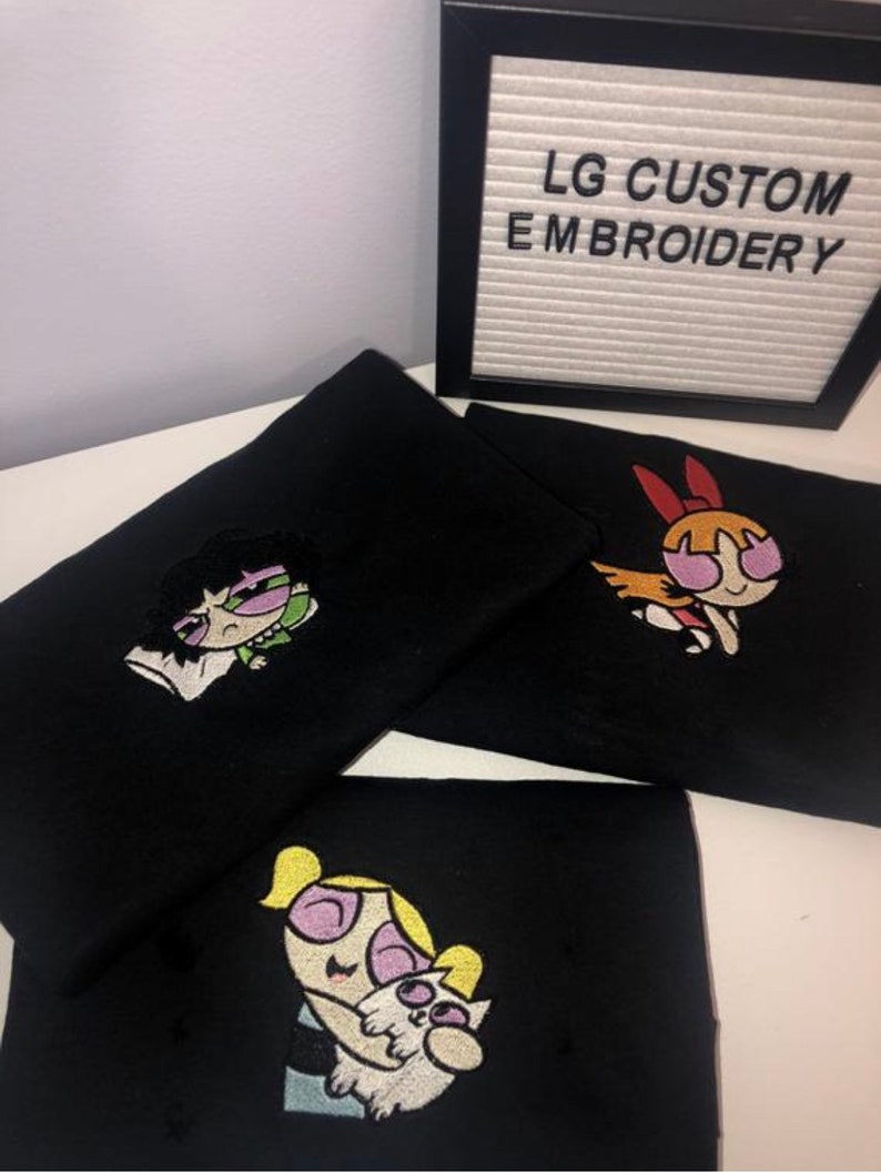 May include: Three black t-shirts with embroidered images of the Powerpuff Girls. The first shirt features Bubbles holding a white cat. The second shirt features Buttercup with a green and purple outfit. The third shirt features Blossom with a red and orange outfit. The shirts are displayed on a white surface with a sign that reads "LG CUSTOM EMBROIDERY".