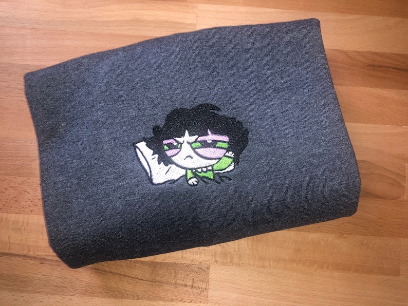 May include: A dark gray sweatshirt with an embroidered cartoon character wearing green and pink. The character has black hair and is holding a pillow.
