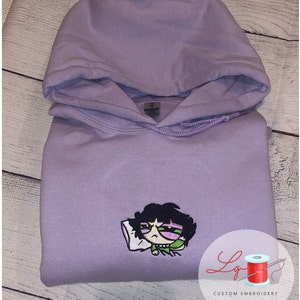 May include: A lavender hoodie with an embroidered image of a cartoon character with black hair, green eyes, and a pink shirt. The character is lying on a white pillow. The hoodie has a drawstring hood and a kangaroo pocket. The text "Lg Custom Embroidery" is visible in the lower right corner of the image.