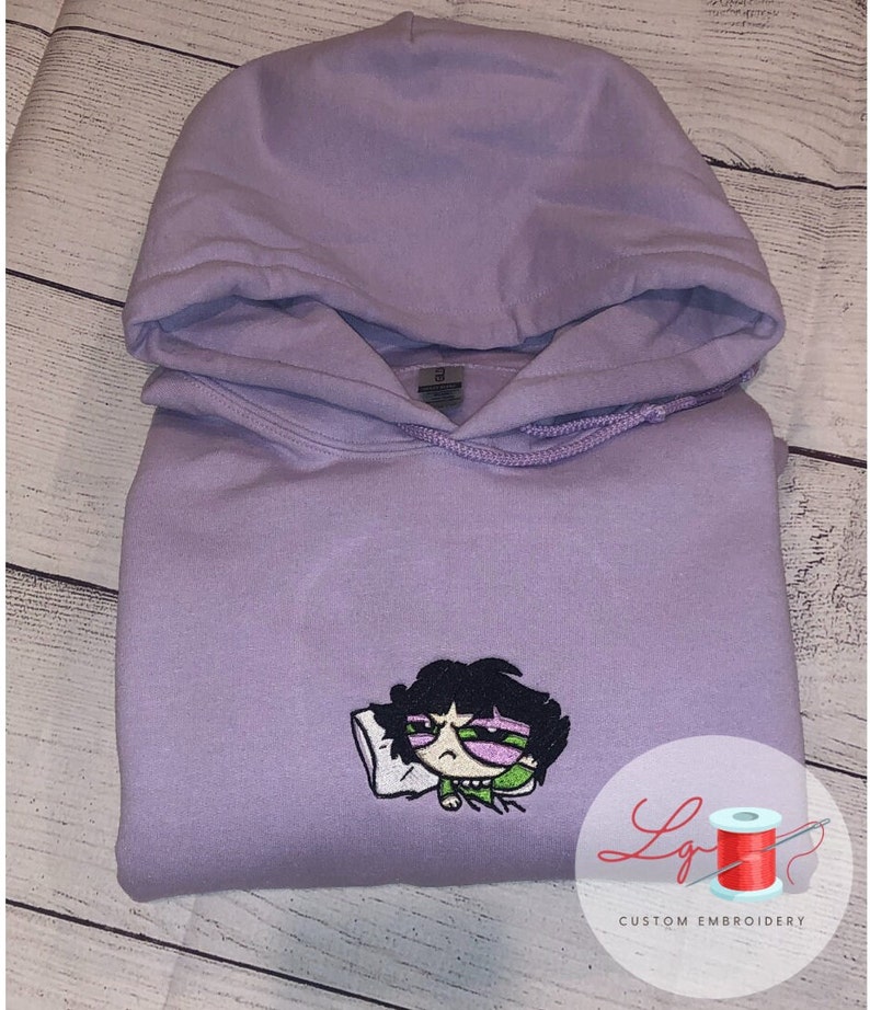 May include: A lavender hoodie with an embroidered image of a cartoon character with black hair, green eyes, and a pink shirt. The character is lying on a white pillow. The hoodie has a drawstring hood and a kangaroo pocket. The text "Lg Custom Embroidery" is visible in the lower right corner of the image.