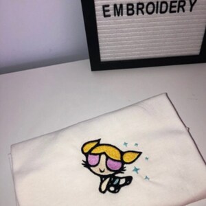 May include: White t-shirt with an embroidered image of a cartoon girl with blonde hair, pink sunglasses, and a blue and white outfit. The text "LG CUSTOM EMBROIDERY" is visible in the background.