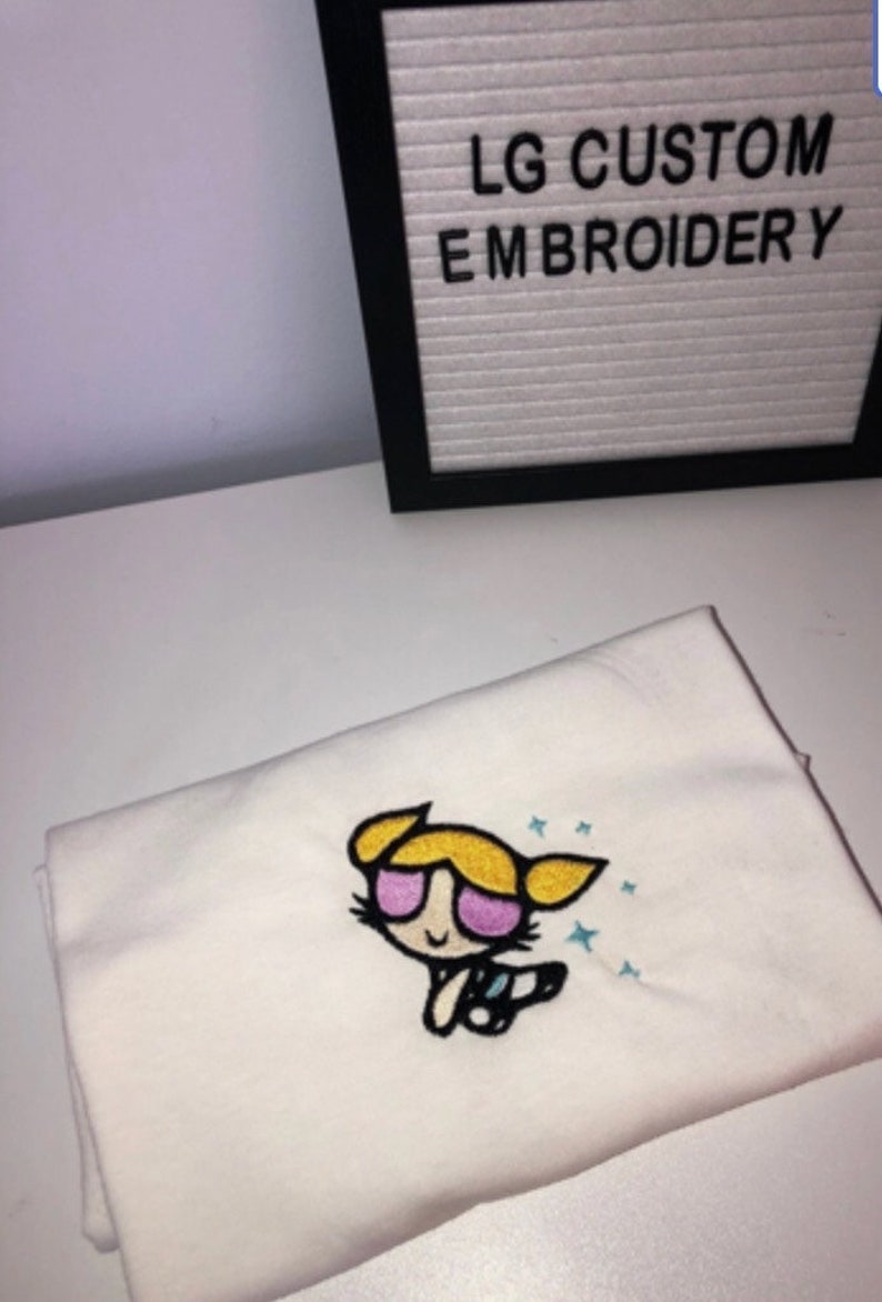 May include: White t-shirt with an embroidered image of a cartoon girl with blonde hair, pink sunglasses, and a blue and white outfit. The text "LG CUSTOM EMBROIDERY" is visible in the background.