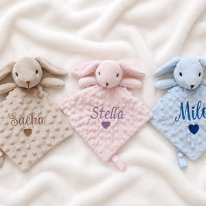Peut inclure: Trois doudous en peluche avec des têtes de lapin en beige, rose clair et bleu clair. Chaque couverture présente un tissu texturé à pois, une petite boucle en tissu et est brodée d'un prénom : Sacha, Stella ou Milo, au-dessus d'un cœur.
