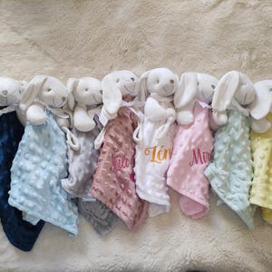 May include: A collection of baby security blankets, each featuring a plush white bunny head and a textured, colorful blanket. The blankets come in various colors, including navy blue, light blue, gray, pink, white, and yellow. Some blankets have embroidered names.