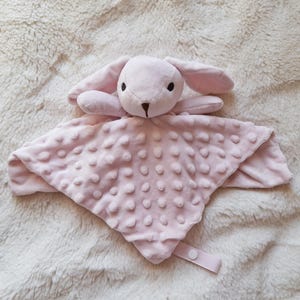 May include: A soft, pink baby security blanket with a plush bunny head and long ears. The blanket has a textured, dotted surface and a ribbon with a button closure. The bunny has black eyes and a brown nose.