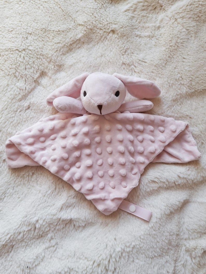 May include: A soft, pink baby security blanket with a plush bunny head and long ears. The blanket has a textured, dotted surface and a ribbon with a button closure. The bunny has black eyes and a brown nose.