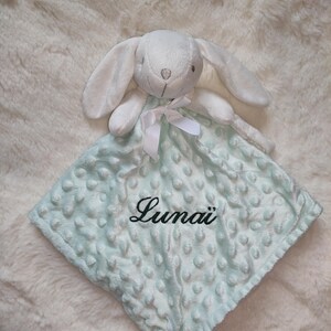 May include: A white plush bunny head attached to a light blue baby blanket. The blanket has a textured, dotted pattern and the name "Luna&iuml;" embroidered in black script. A white ribbon is tied around the bunny's neck.