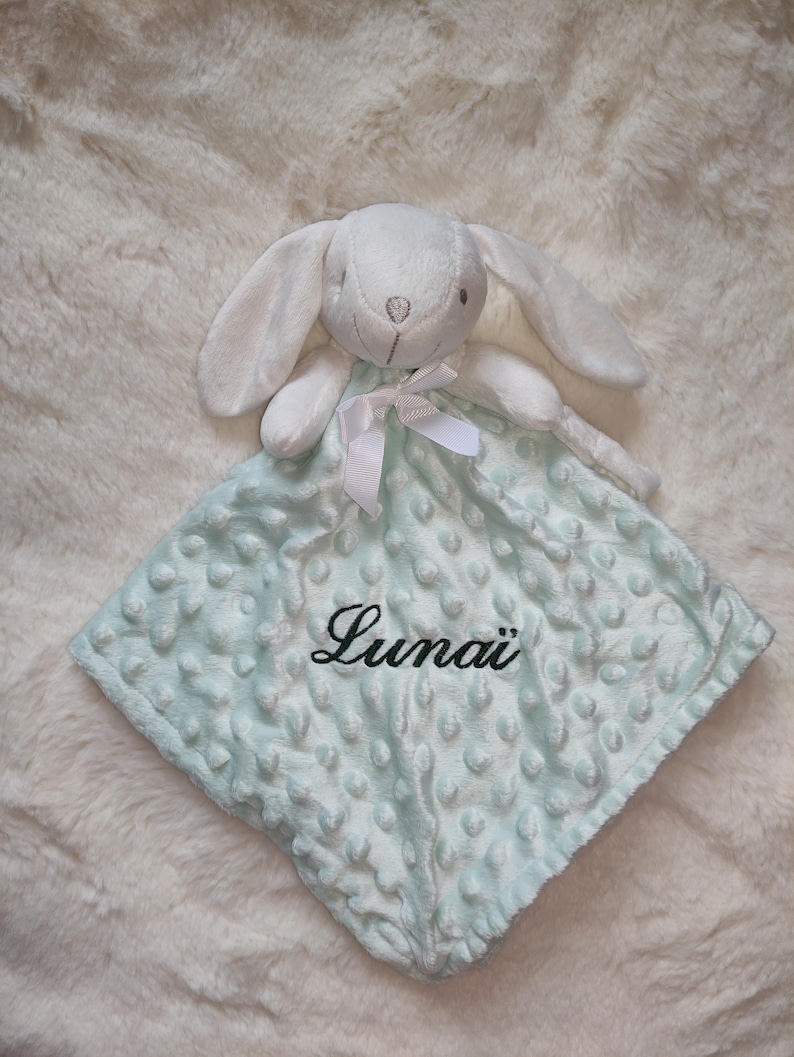 May include: A white plush bunny head attached to a light blue baby blanket. The blanket has a textured, dotted pattern and the name "Luna&iuml;" embroidered in black script. A white ribbon is tied around the bunny's neck.