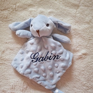 May include: A light blue baby security blanket with a plush bunny head and the name "Gabin" embroidered in navy blue. The blanket has a textured, dotted pattern and a light blue ribbon with a snap closure.