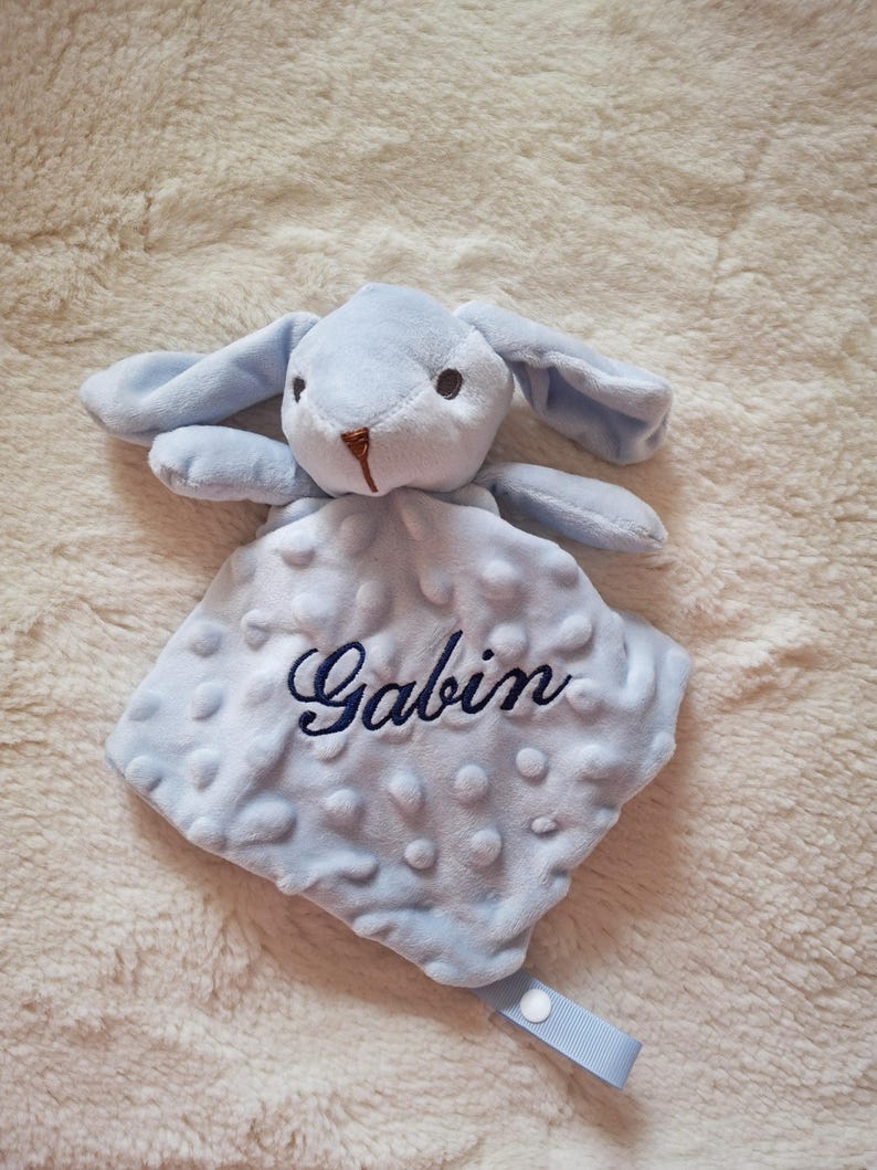 May include: A light blue baby security blanket with a plush bunny head and the name "Gabin" embroidered in navy blue. The blanket has a textured, dotted pattern and a light blue ribbon with a snap closure.