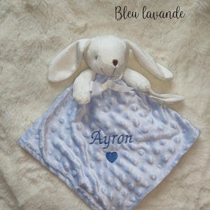 May include: A white plush bunny toy with long ears and a bow, resting on a light blue, square baby blanket. The blanket has a textured surface and the name "Ayron" is embroidered on it, along with a blue heart. The text "Bleu lavande" is written in cursive.