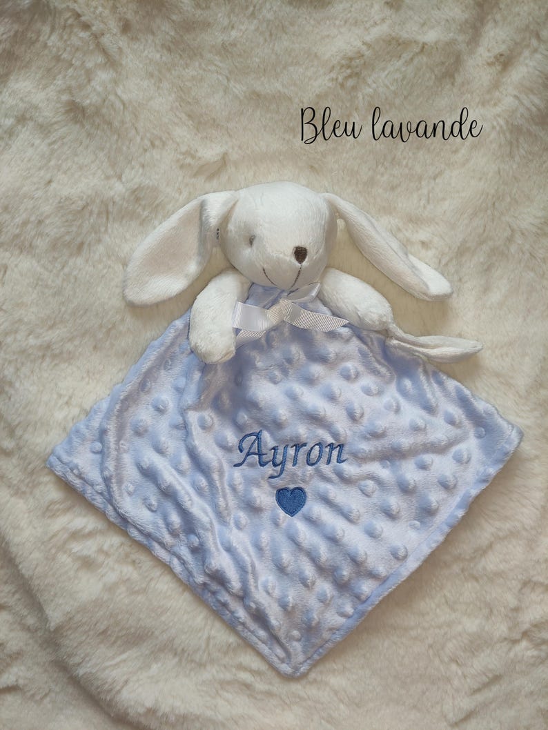 May include: A white plush bunny toy with long ears and a bow, resting on a light blue, square baby blanket. The blanket has a textured surface and the name "Ayron" is embroidered on it, along with a blue heart. The text "Bleu lavande" is written in cursive.