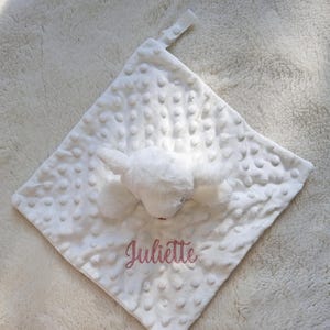 May include: White baby security blanket with a plush bunny head and the name "Juliette" embroidered in pink. The blanket has a textured, dotted surface and a small loop for attachment.