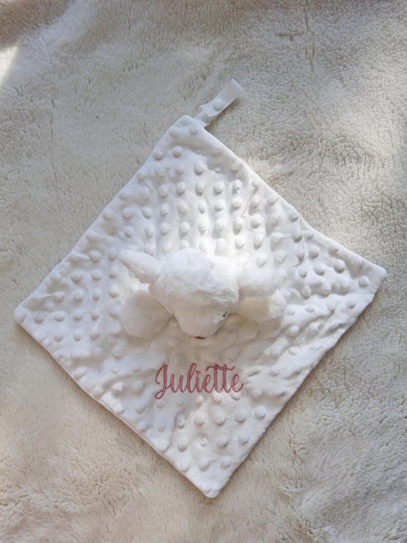 May include: White baby security blanket with a plush bunny head and the name "Juliette" embroidered in pink. The blanket has a textured, dotted surface and a small loop for attachment.
