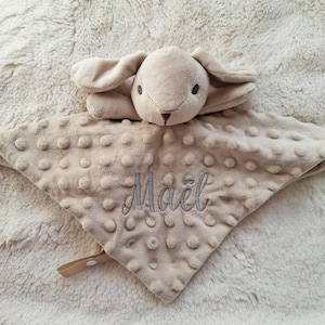 May include: A beige baby security blanket with a plush bunny head and a textured, dotted surface. The blanket has the name "Ma&euml;l" embroidered in gray thread. A small, tan strap with a snap closure is attached.
