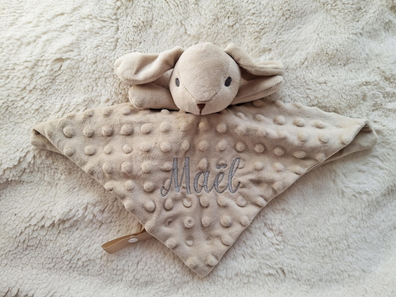 May include: A beige baby security blanket with a plush bunny head and a textured, dotted surface. The blanket has the name "Ma&euml;l" embroidered in gray thread. A small, tan strap with a snap closure is attached.