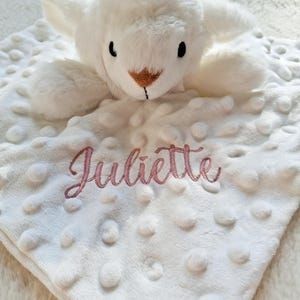 May include: A white plush baby blanket with a soft, textured surface and a stuffed lamb head. The name "Juliette" is embroidered in pink script on the blanket. The lamb has black eyes and a brown nose.