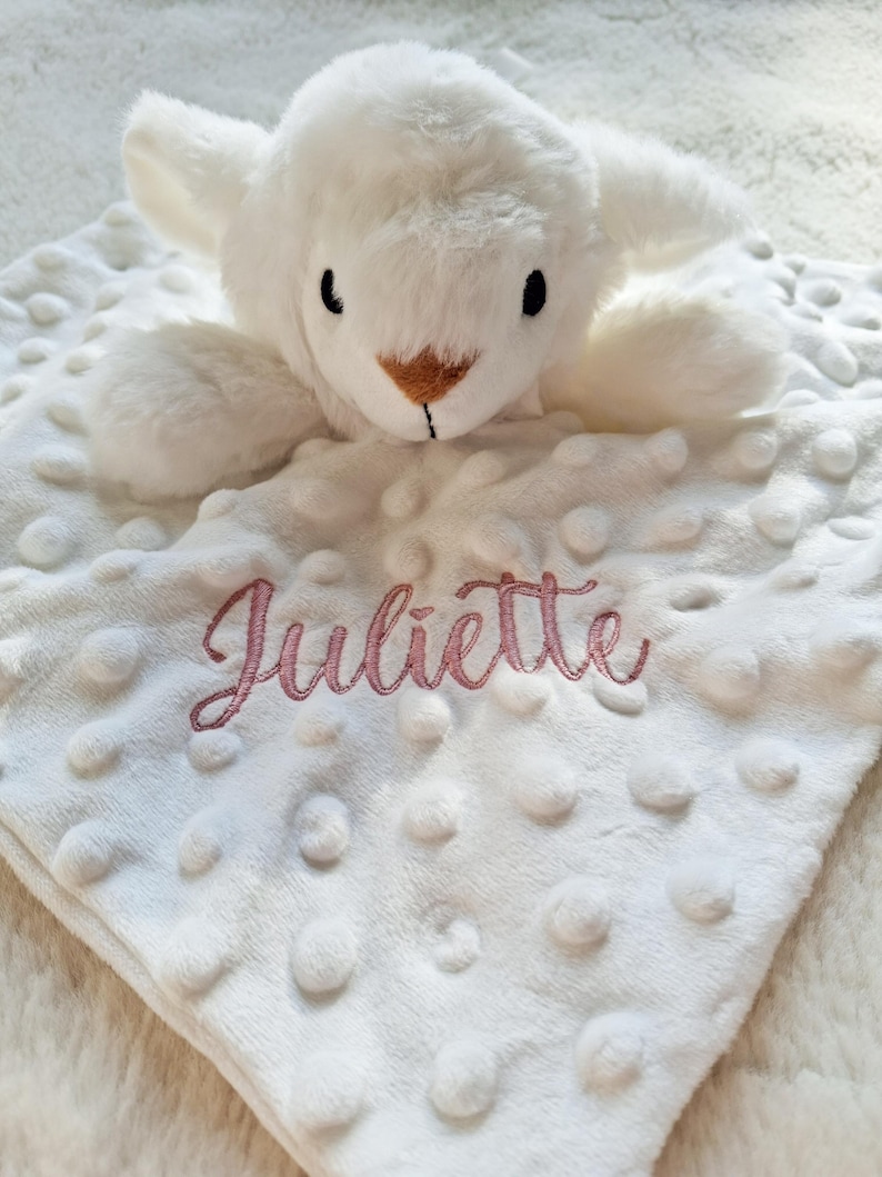 May include: A white plush baby blanket with a soft, textured surface and a stuffed lamb head. The name "Juliette" is embroidered in pink script on the blanket. The lamb has black eyes and a brown nose.