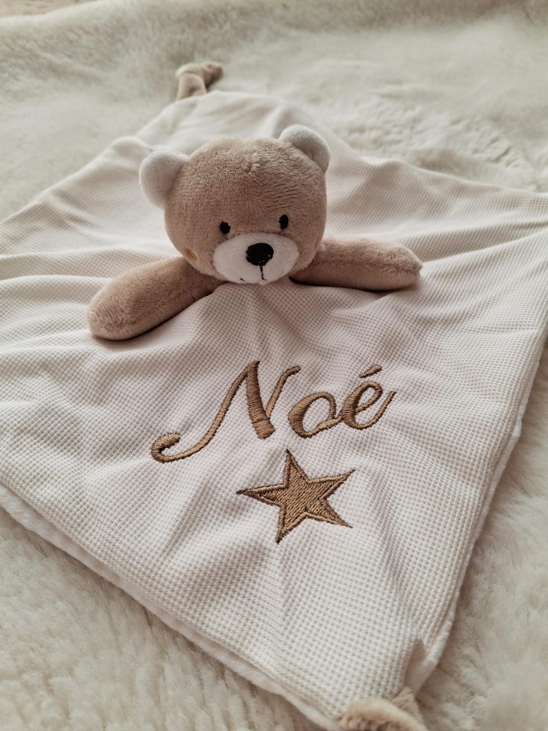 May include: A beige baby security blanket with a teddy bear head and arms attached. The blanket has the embroidered name "No&eacute;" and a star. The blanket is made of a waffle-weave fabric.