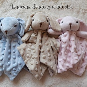 May include: Three plush bunny security blankets in pastel colors: light blue, tan, and pink. Each bunny has long ears, a stitched face, and a textured blanket body. The text "Nouveaux doudous &agrave; adopter" is at the top.