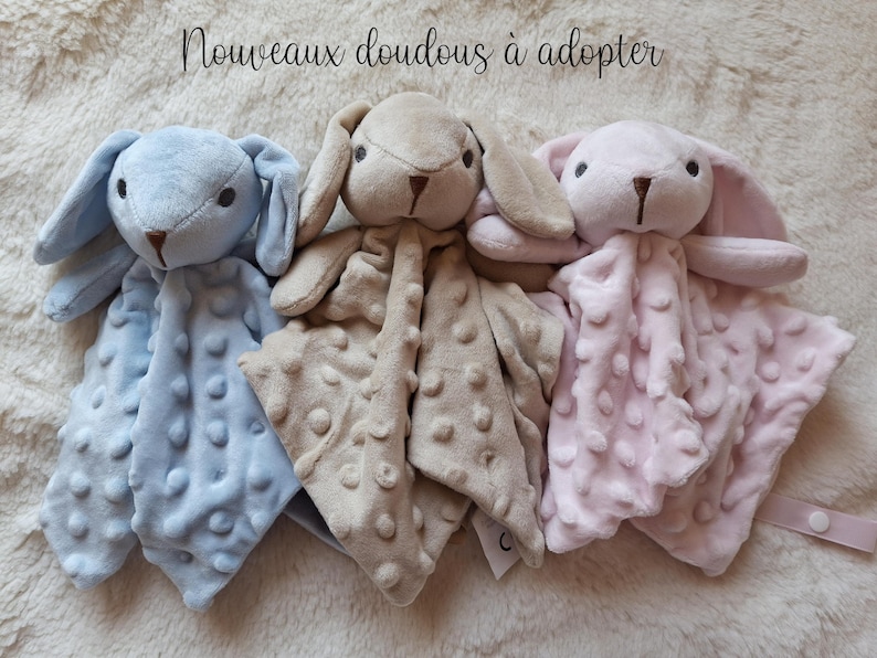 May include: Three plush bunny security blankets in pastel colors: light blue, tan, and pink. Each bunny has long ears, a stitched face, and a textured blanket body. The text "Nouveaux doudous &agrave; adopter" is at the top.