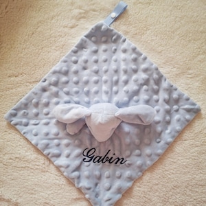 May include: A light blue baby security blanket with a textured, dotted surface. The blanket features a white plush bunny head and the name "Gabin" embroidered in black script. A light blue ribbon loop is attached to the corner.