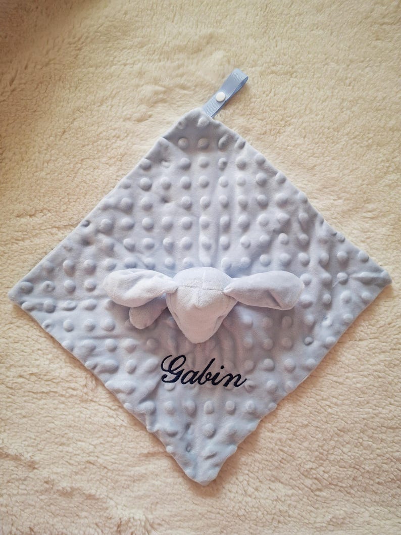 May include: A light blue baby security blanket with a textured, dotted surface. The blanket features a white plush bunny head and the name "Gabin" embroidered in black script. A light blue ribbon loop is attached to the corner.