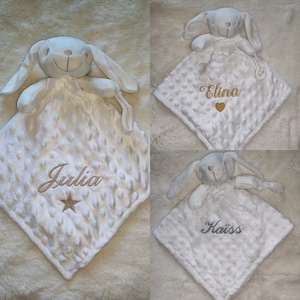 May include: White baby security blanket with a plush bunny head and a textured, dotted surface. The blanket has embroidered names and a star or heart design. The bunny has long ears and a ribbon bow.