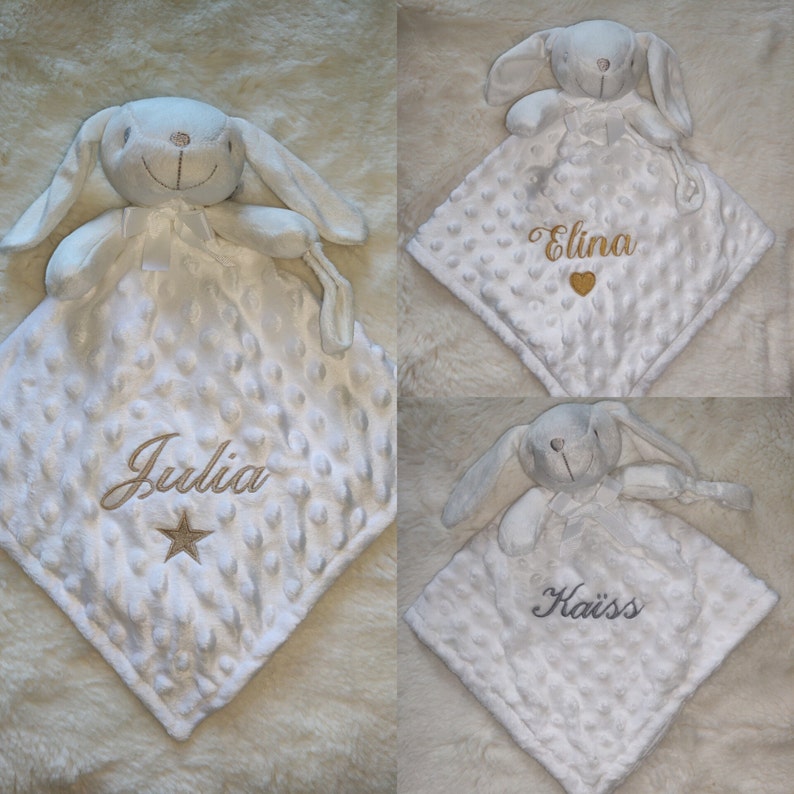 May include: White baby security blanket with a plush bunny head and a textured, dotted surface. The blanket has embroidered names and a star or heart design. The bunny has long ears and a ribbon bow.