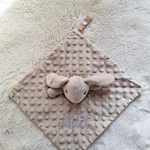 May include: A beige baby security blanket with a plush bunny head and ears attached. The blanket has a textured, dotted surface and the name "Ma&euml;l" is embroidered in light blue thread. A fabric loop with a button is attached.