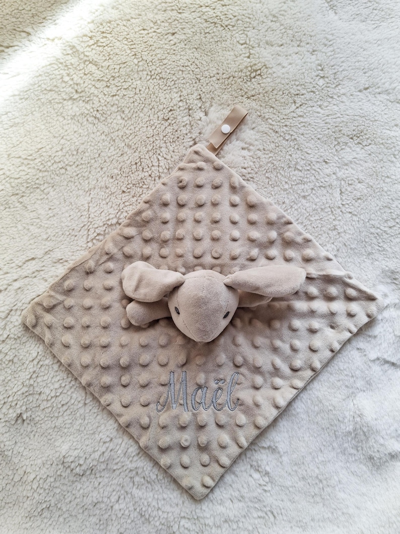 May include: A beige baby security blanket with a plush bunny head and ears attached. The blanket has a textured, dotted surface and the name "Ma&euml;l" is embroidered in light blue thread. A fabric loop with a button is attached.