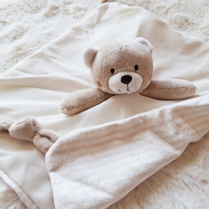 May include: A beige baby security blanket with a teddy bear head and arms attached. The blanket has a waffle weave texture on one side and a soft, striped fabric on the other. The bear has a brown nose and embroidered eyes.
