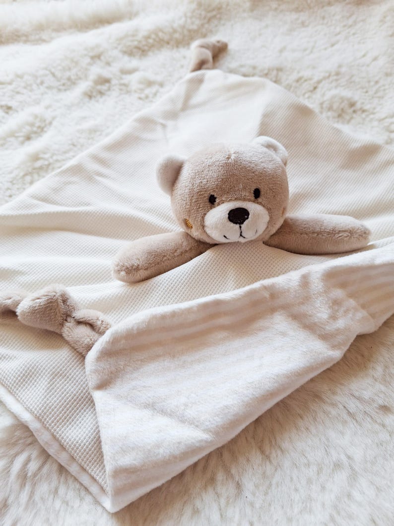 May include: A beige baby security blanket with a teddy bear head and arms attached. The blanket has a waffle weave texture on one side and a soft, striped fabric on the other. The bear has a brown nose and embroidered eyes.