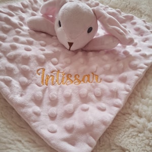 May include: A pink baby security blanket with a plush bunny head and ears. The blanket has a textured surface with raised dots and the name "Intissar" embroidered in gold thread.