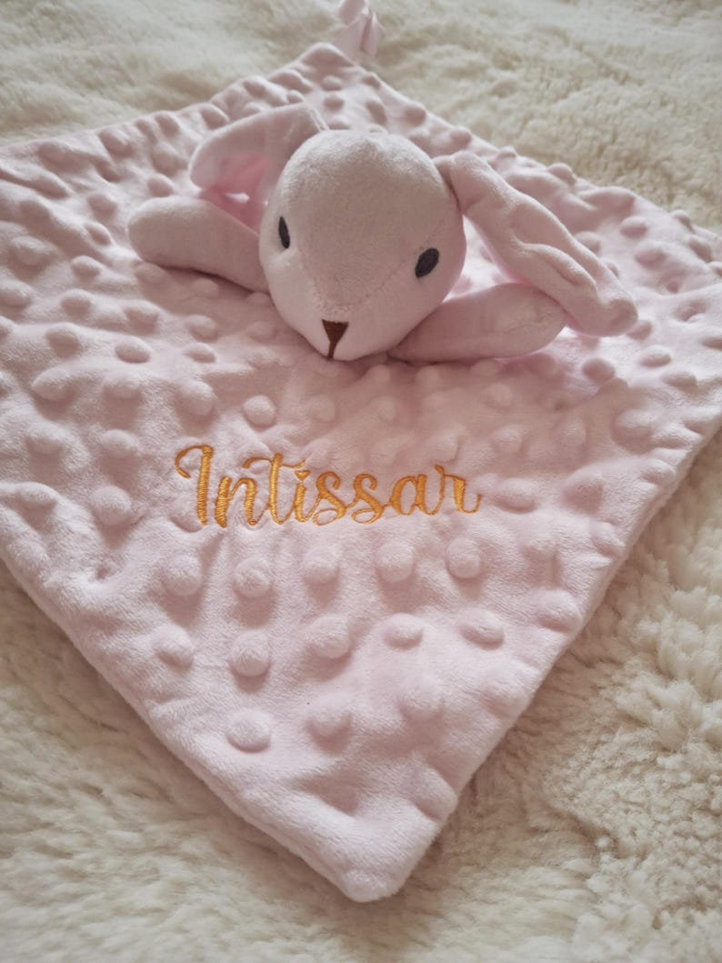 May include: A pink baby security blanket with a plush bunny head and ears. The blanket has a textured surface with raised dots and the name "Intissar" embroidered in gold thread.