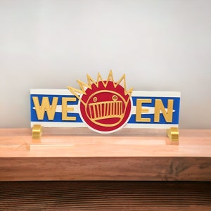 May include: A white sign with blue and red stripes and the words "WE EN" in gold letters. A red and gold cartoon face with a crown of spikes is in the center of the sign.