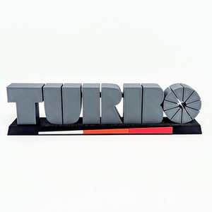 Peut inclure: Le mot "TURBO" imprimé en 3D en gris avec un design circulaire segmenté gris derrière. Le mot est sur une base noire avec des rayures rouges et blanches.