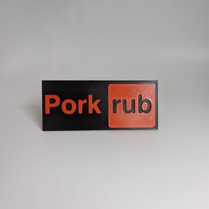 May include: A black and orange rectangular sign with the text "Pork rub" printed in orange.