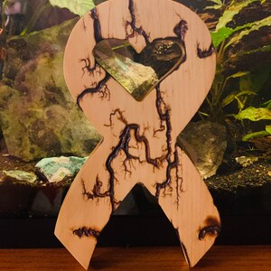 May include: A wooden ribbon with a heart-shaped cutout in the center. The ribbon is decorated with a wood-burned design that resembles lightning bolts. The ribbon is light brown in color.