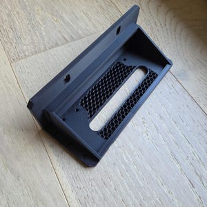 May include: A black, 3D-printed bracket with a rectangular shape and a honeycomb pattern. The bracket has a lip on one side and several holes for mounting. The object is made of plastic and appears to be a component for electronics or a similar device.