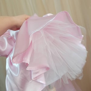 May include: A light pink satin dress featuring a voluminous skirt with multiple layers of white tulle underneath. The fabric has a smooth, reflective finish, and the skirt is gathered to show the structured, layered construction of the hemline.