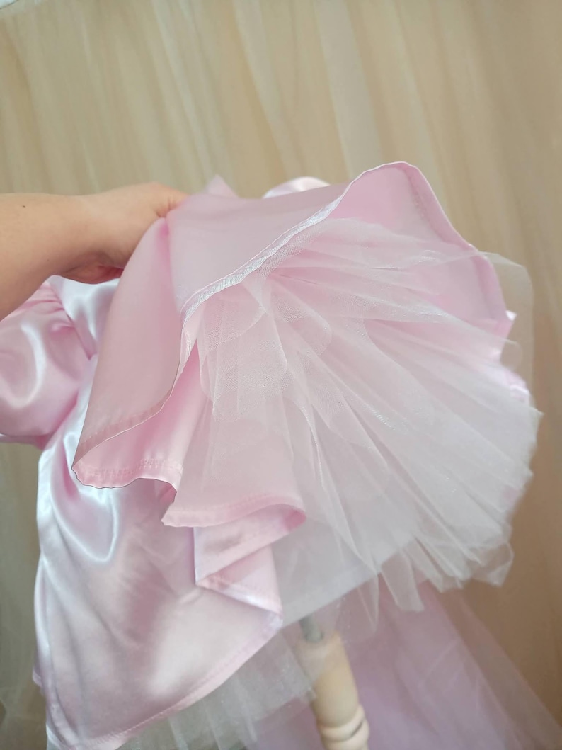 May include: A light pink satin dress featuring a voluminous skirt with multiple layers of white tulle underneath. The fabric has a smooth, reflective finish, and the skirt is gathered to show the structured, layered construction of the hemline.