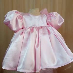 May include: A light pink satin dress featuring voluminous puff sleeves, a round neckline, and a full, gathered skirt. The garment has a smooth, shiny finish and a structured silhouette, displayed on a mannequin to showcase the flared design.