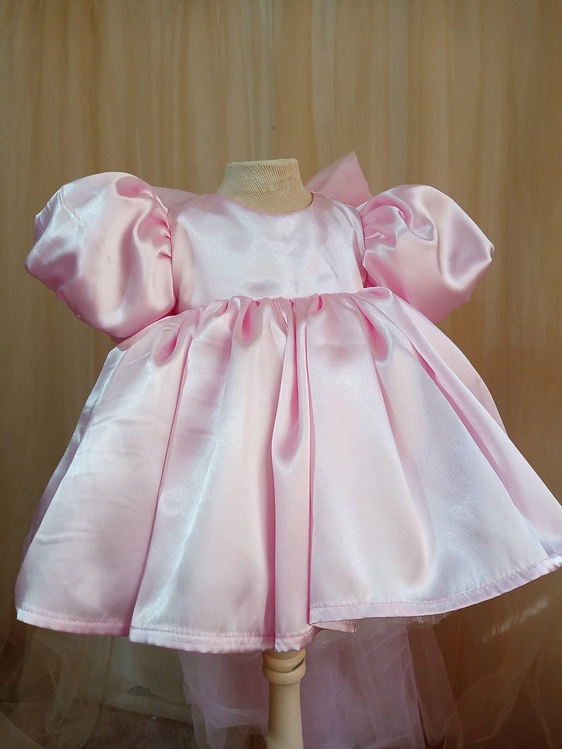 May include: A light pink satin dress featuring voluminous puff sleeves, a round neckline, and a full, gathered skirt. The garment has a smooth, shiny finish and a structured silhouette, displayed on a mannequin to showcase the flared design.